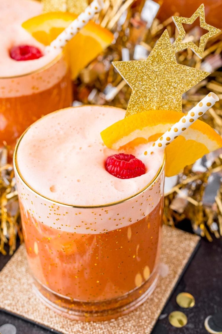 A close up photo of party punch garnished with a raspberry and orange slice on a gold coaster with party decorations in the background.