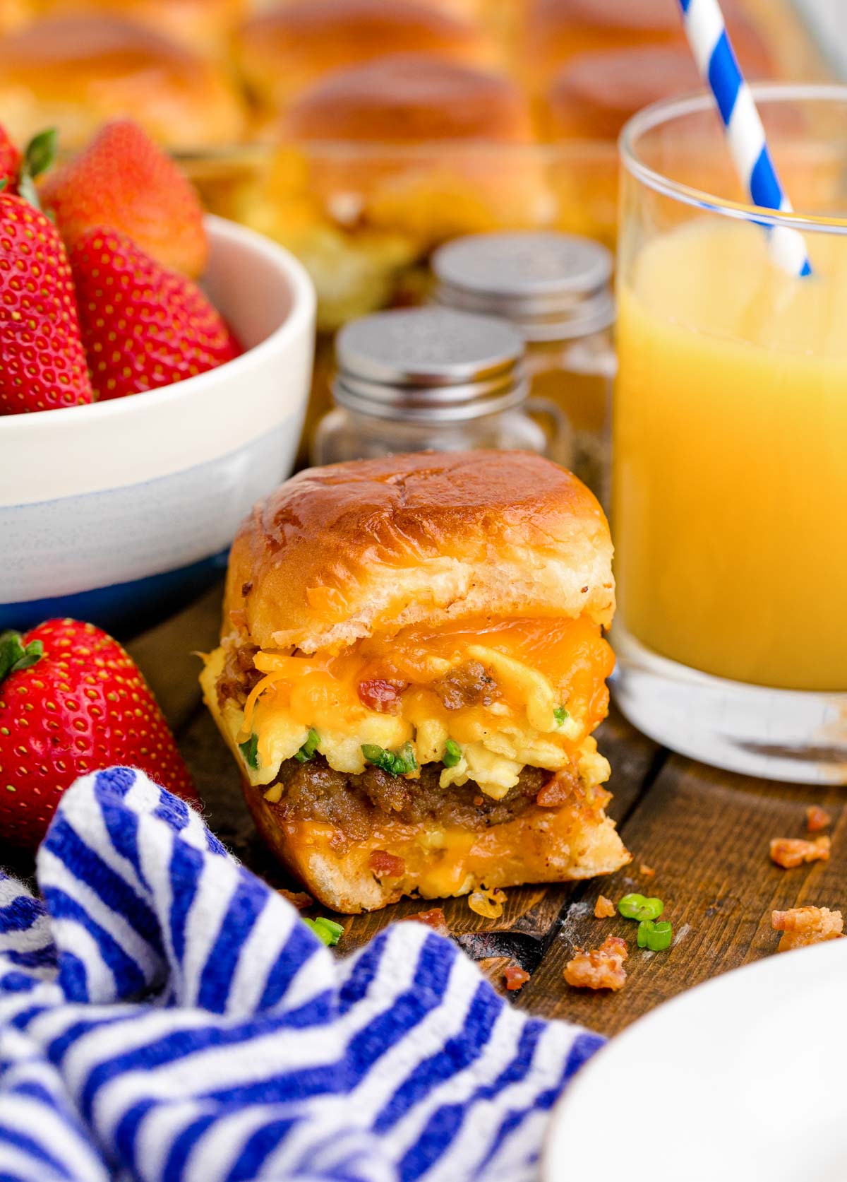 Close up photo of breakfast sliders on a wooden table with a glass of orange juice and bowl of strawberries around it.