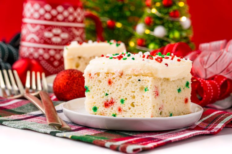 A slice of funfetti Christmas cake on a white plate on a holiday napkin with decor in the background.