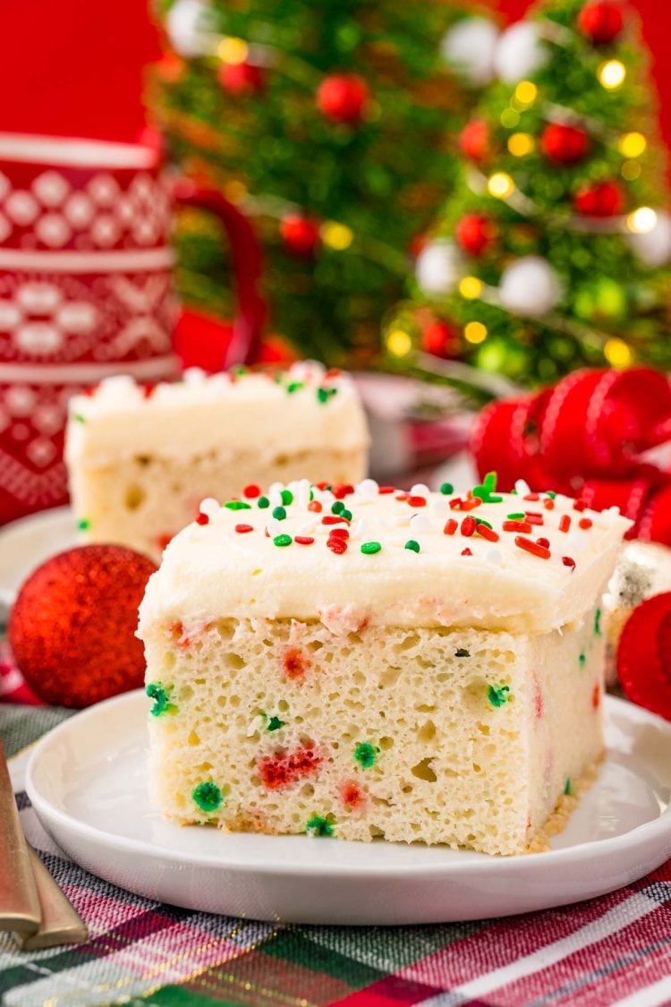 A slice of funfetti Christmas cake on a white plate on a holiday napkin with decor in the background.