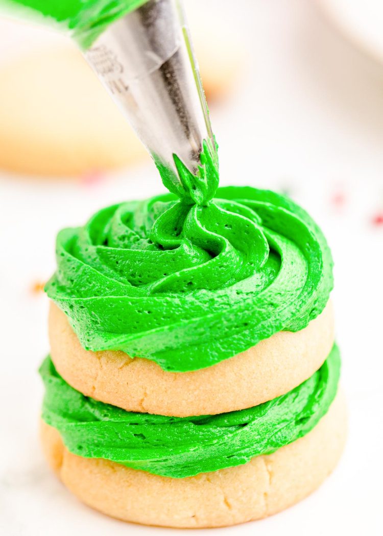 Close up photo of sugar cookies stacked with green frosting in between. The top cookie is having frosting piped on.