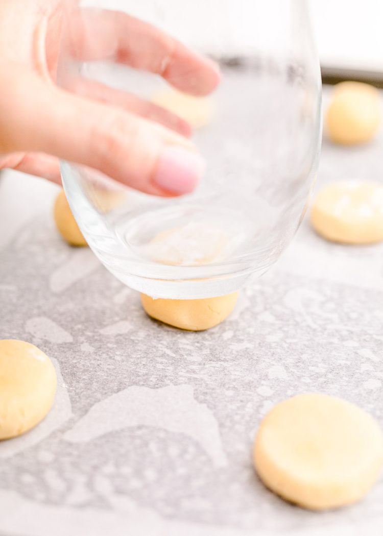 A woman's hand using the bottom of a glass to press balls of sugar cookie dough into flat discs.