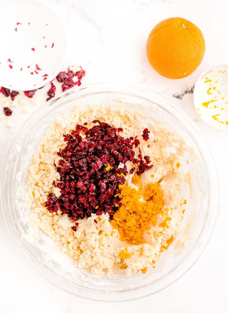 Orange zest and craisins added to a large glass mixing bowl with cookie dough in it.