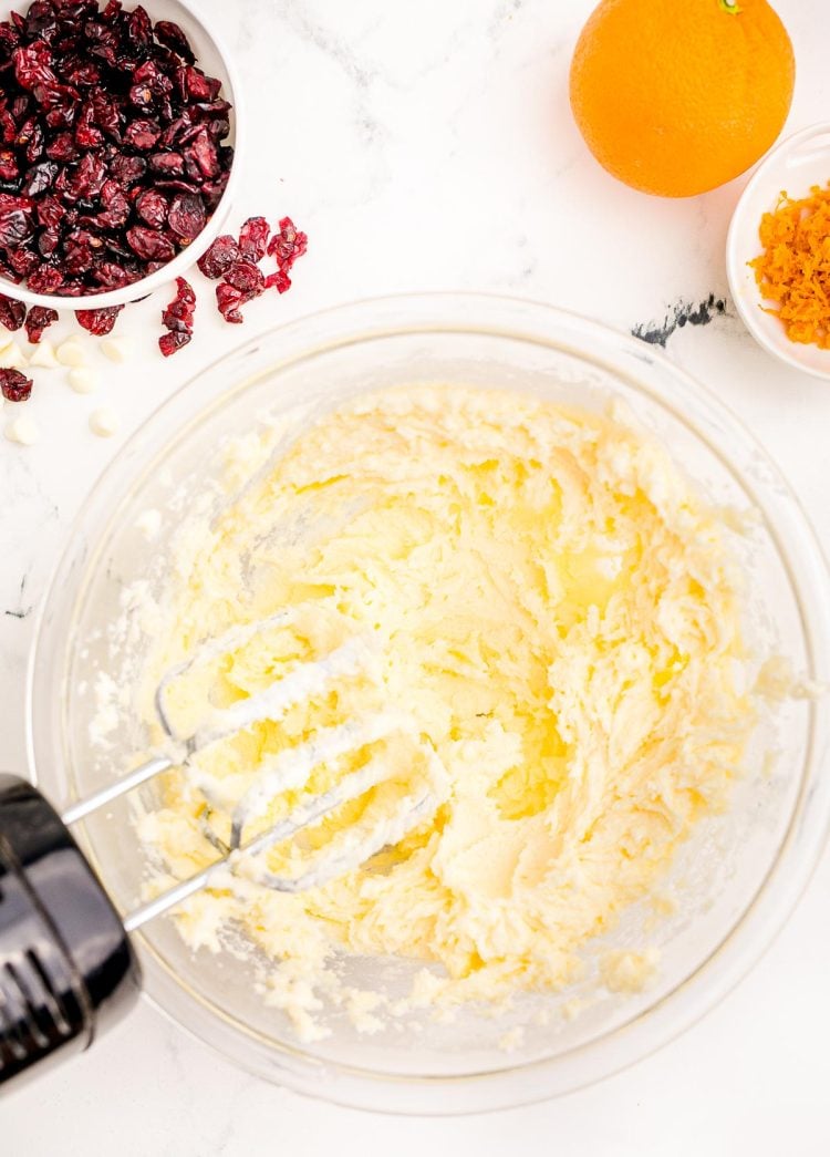 Butter and sugar being creamed together in a large glass mixing bowl. Craisins and orange on the side.