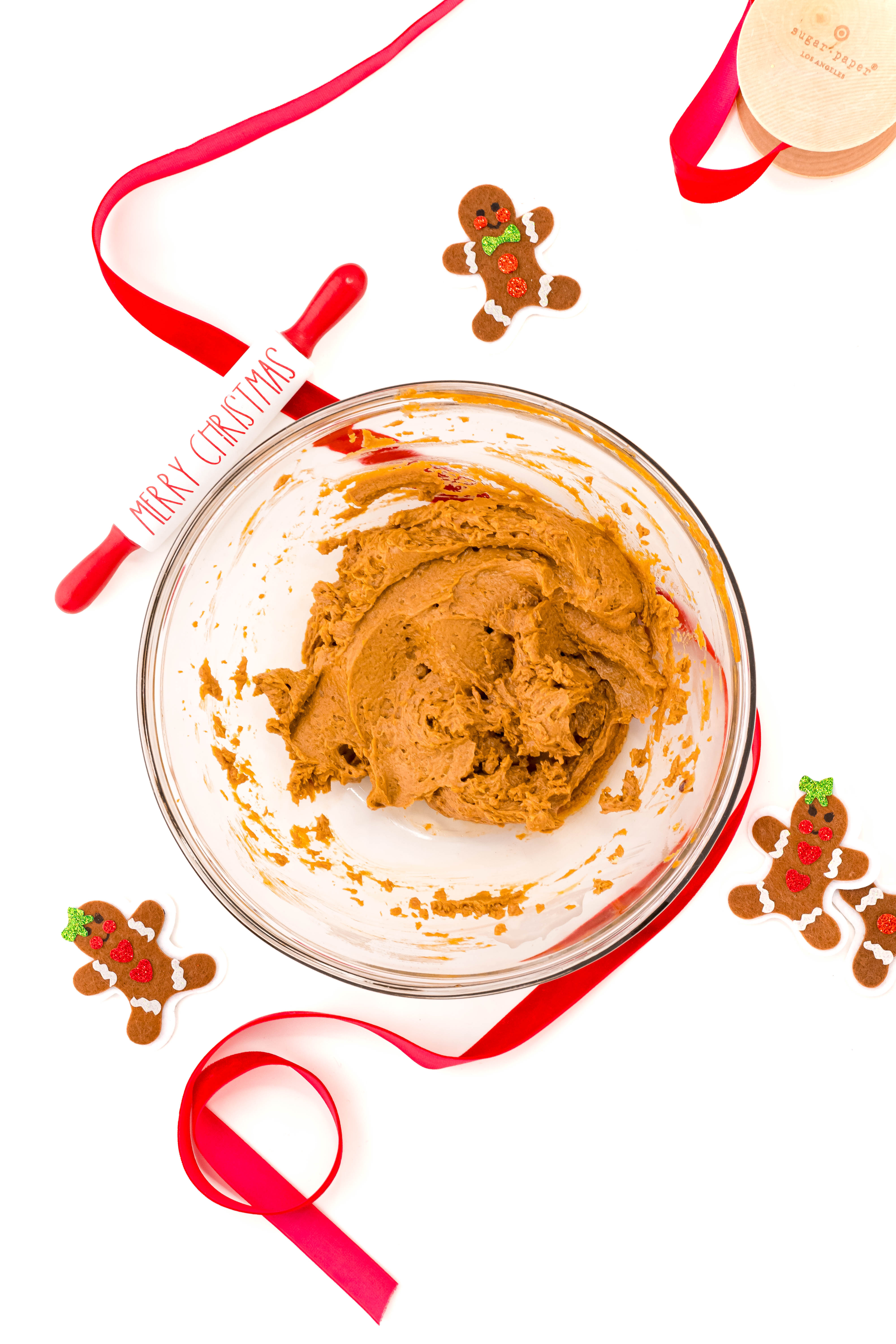 Overhead photo of a glass bowl filled with edible gingerbread cookie dough with red ribbon and gingerbread men around it.