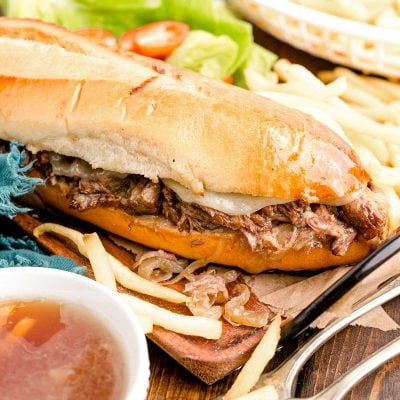 Close up photo of french dip sandwiches on a wooden board with french fries.