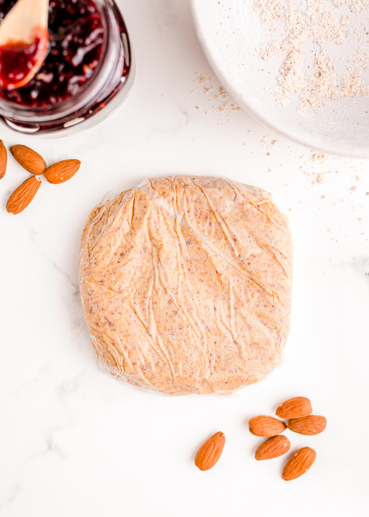 Linzer cookie dough wrapped in plastic wrapped on a table with almonds and raspberry jam around them.