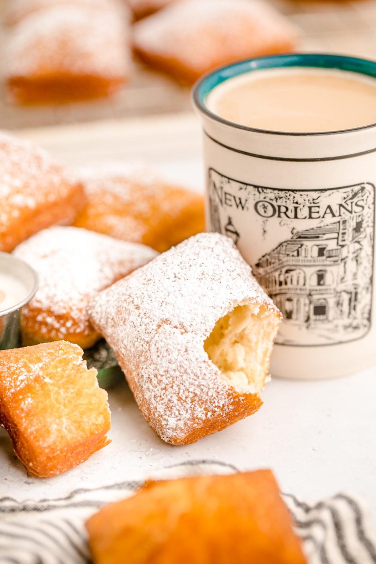 A Beignets covered in powdered sugar leaning on a mug of coffee with a bite taken out of it.