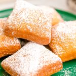 Close up photo of beignets on a green plate with a mug of coffee and more beignets in the background.