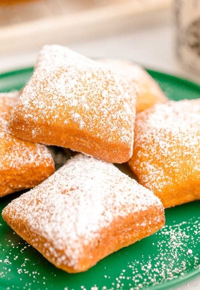 Close up photo of beignets on a green plate with a mug of coffee and more beignets in the background.