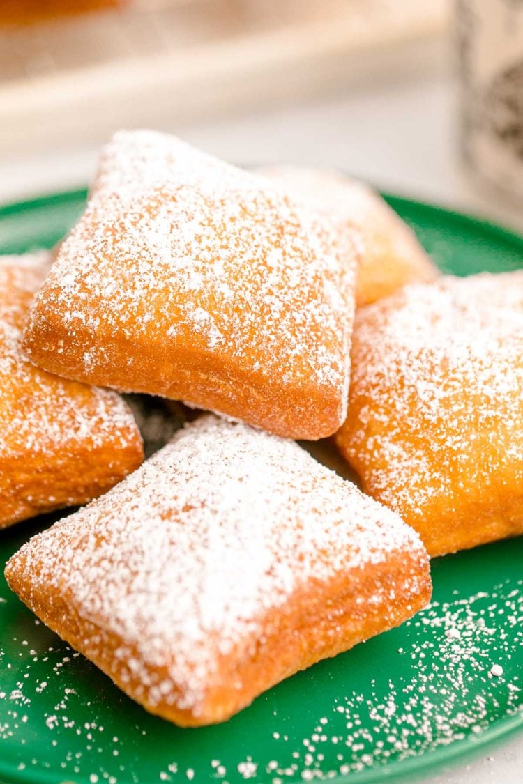 Close up photo of beignets on a green plate with a mug of coffee and more beignets in the background.