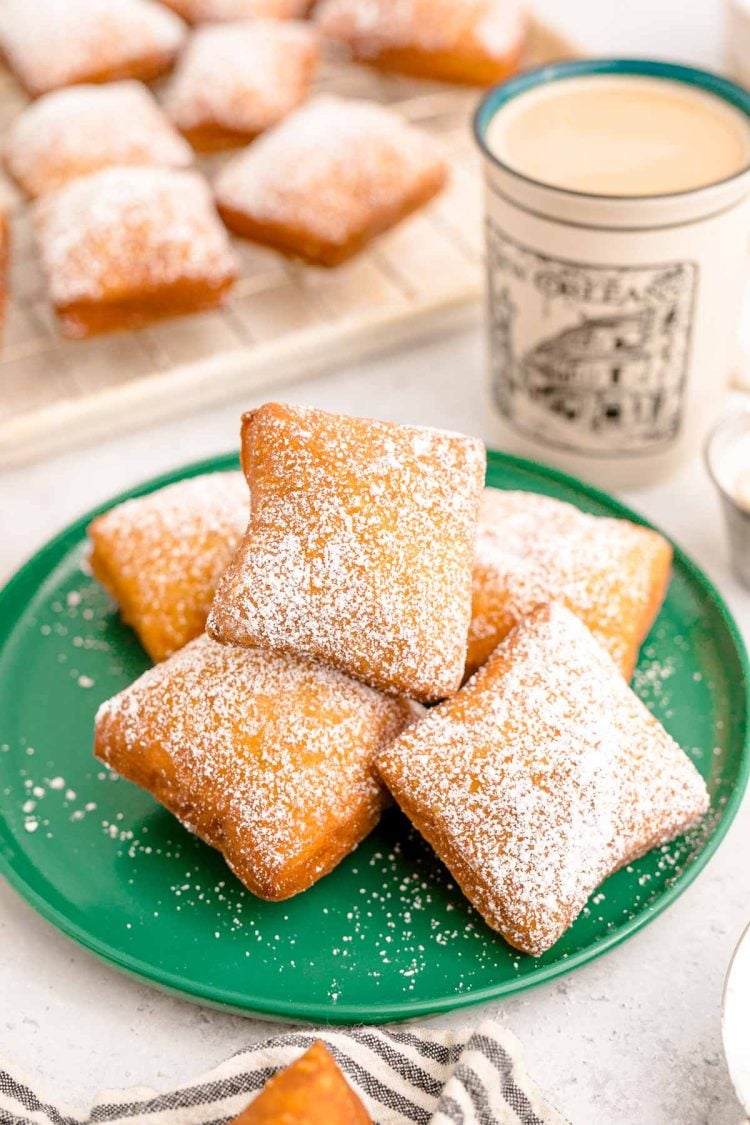 Close up photo of beignets on a green plate with a mug of coffee and more beignets in the background.
