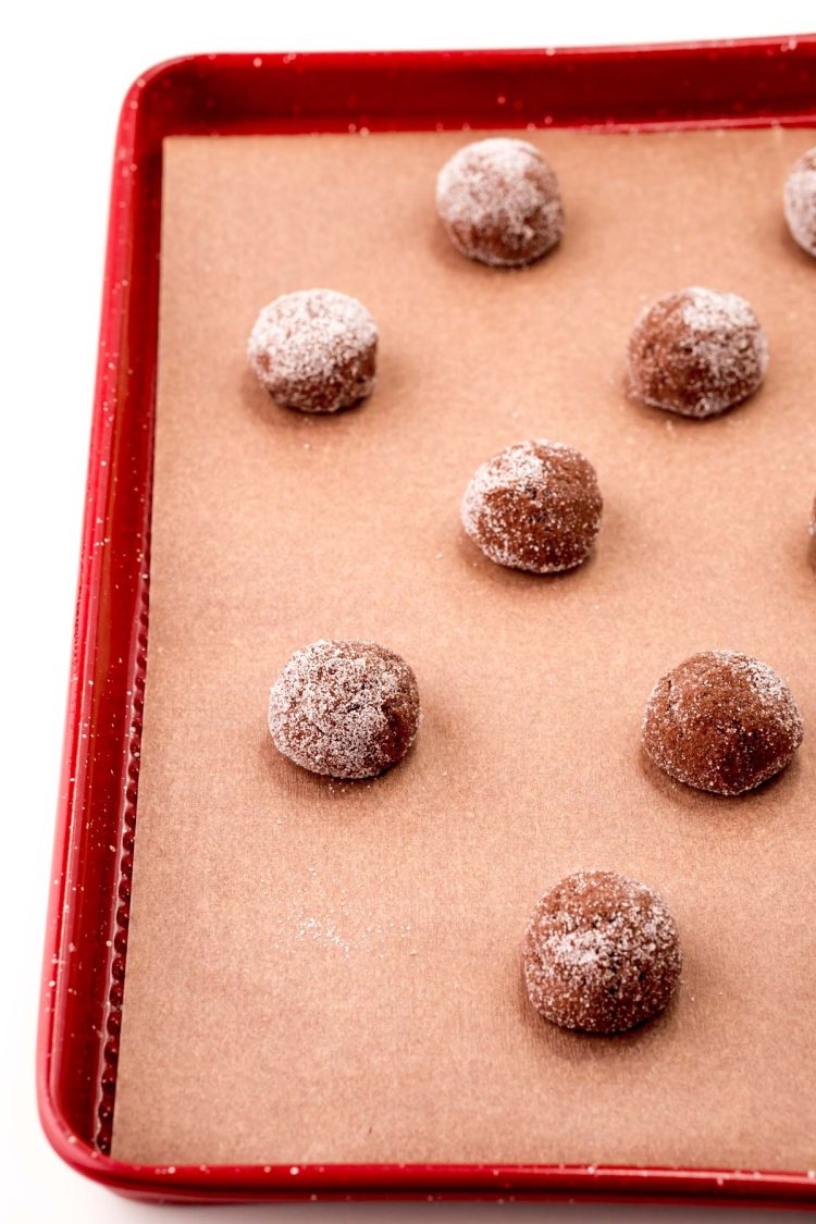 Balls of chocolate cookie dough rolled in granulated sugar on a red parchment-lined baking sheet.