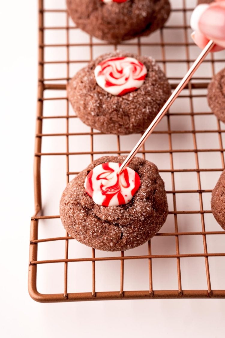 Chocolate thumbprint cookies on a wire rack with a woman's hand swirling the melted candy with a toothpick.