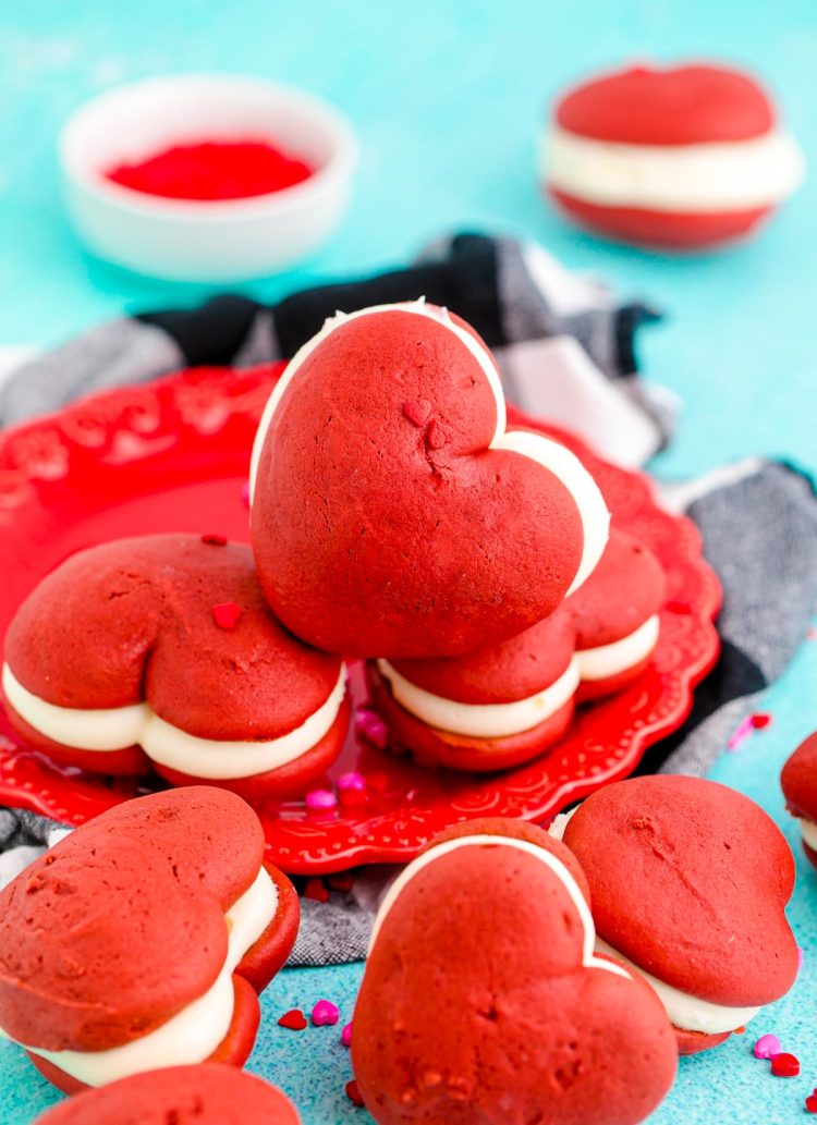 Close up phot of heart shaped whoopie pies on a red plate on a black and white checkered napkin on a blue surface.