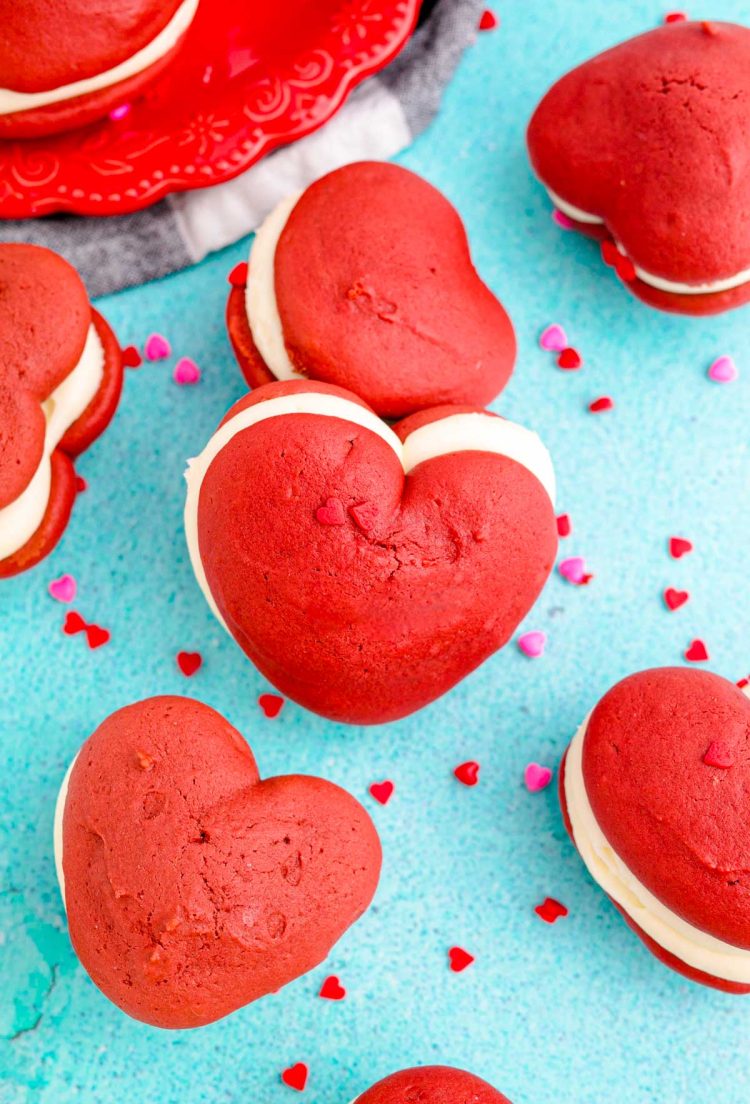 Heart-shaped red velvet whoopie pies on a blue surface surrounded by sprinkles.