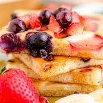 Close up photo of a stack of sheetpan pancakes on a plate with fruit around it.