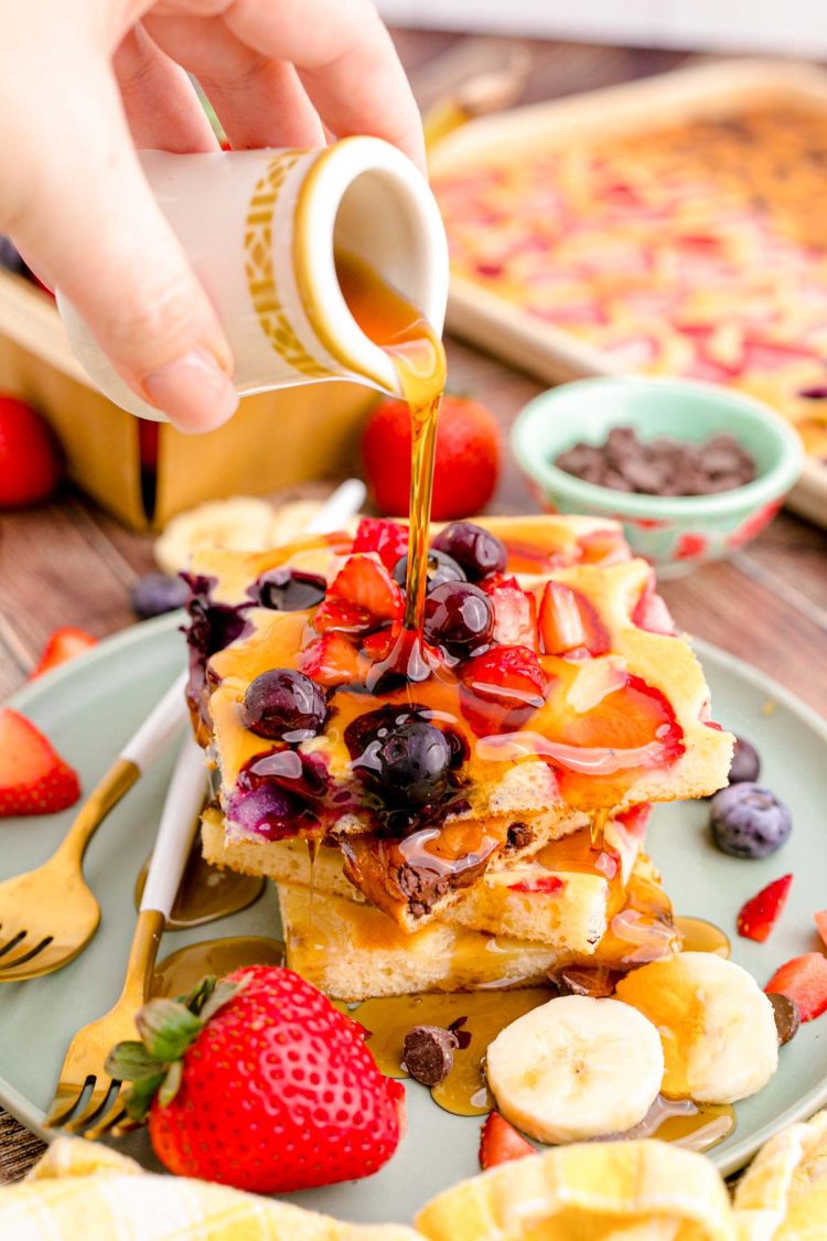 A woman's hand pouring maple syrup over a plate of sheet pan pancakes.