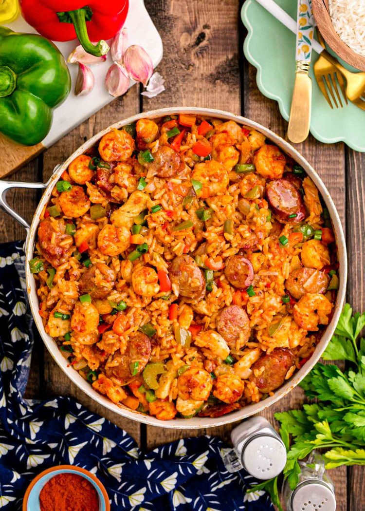 Overhead photo of a pan of jambalaya on a wooden table with a navy napkin and ingredients around it.