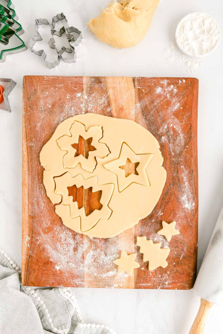 Sugar cookies dough rolled out on a wooden cutting board with holiday shapes cut out of the dough.
