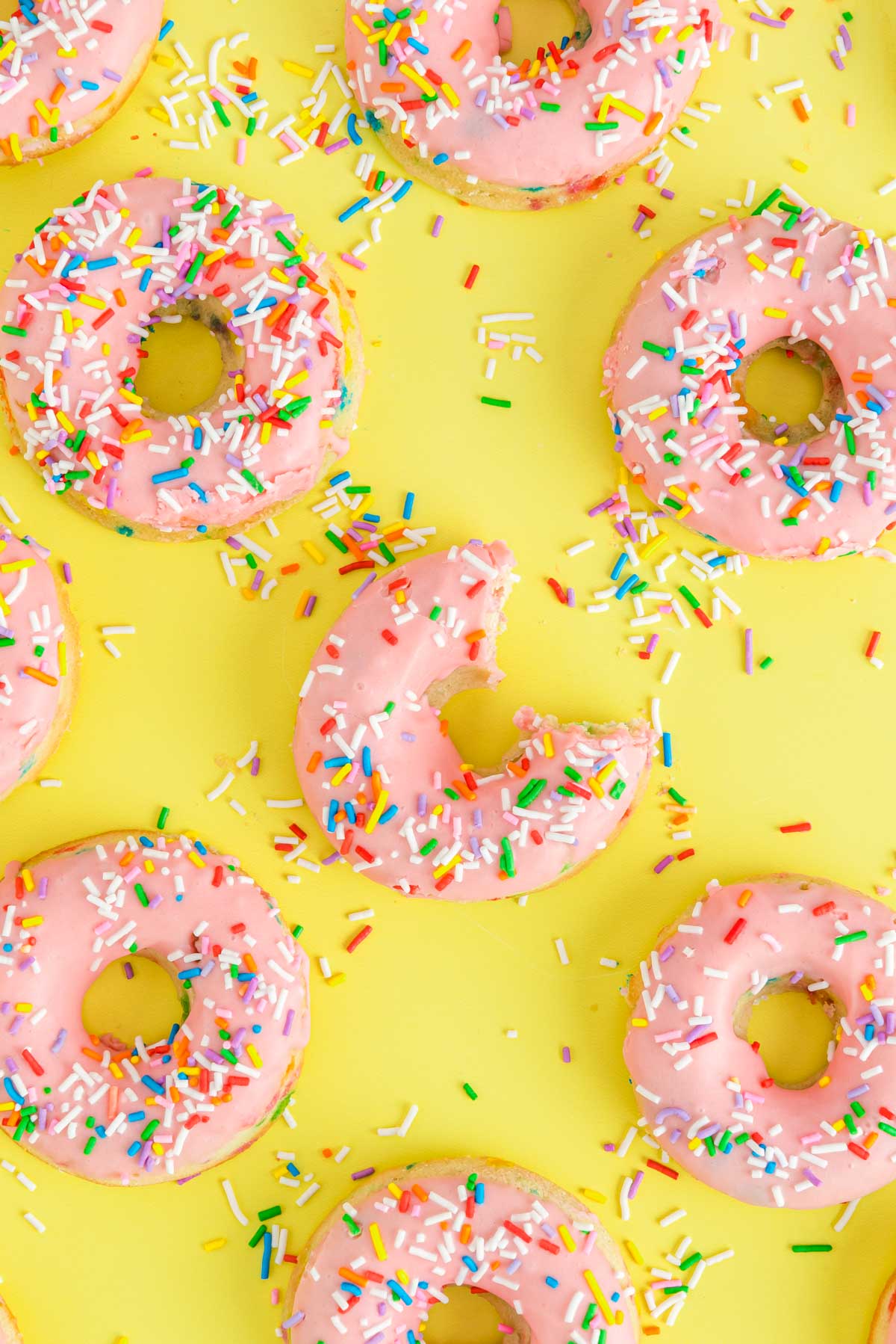 A bite taken out of a birthday cake donut on a yellow surface with more donuts.