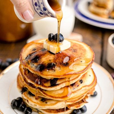 Close up photo of a woman's hand pouring maple syrup over a stack of blueberry pancakes on a white plate on a wooden table.