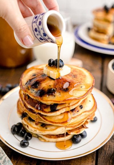 Close up photo of a woman's hand pouring maple syrup over a stack of blueberry pancakes on a white plate on a wooden table.