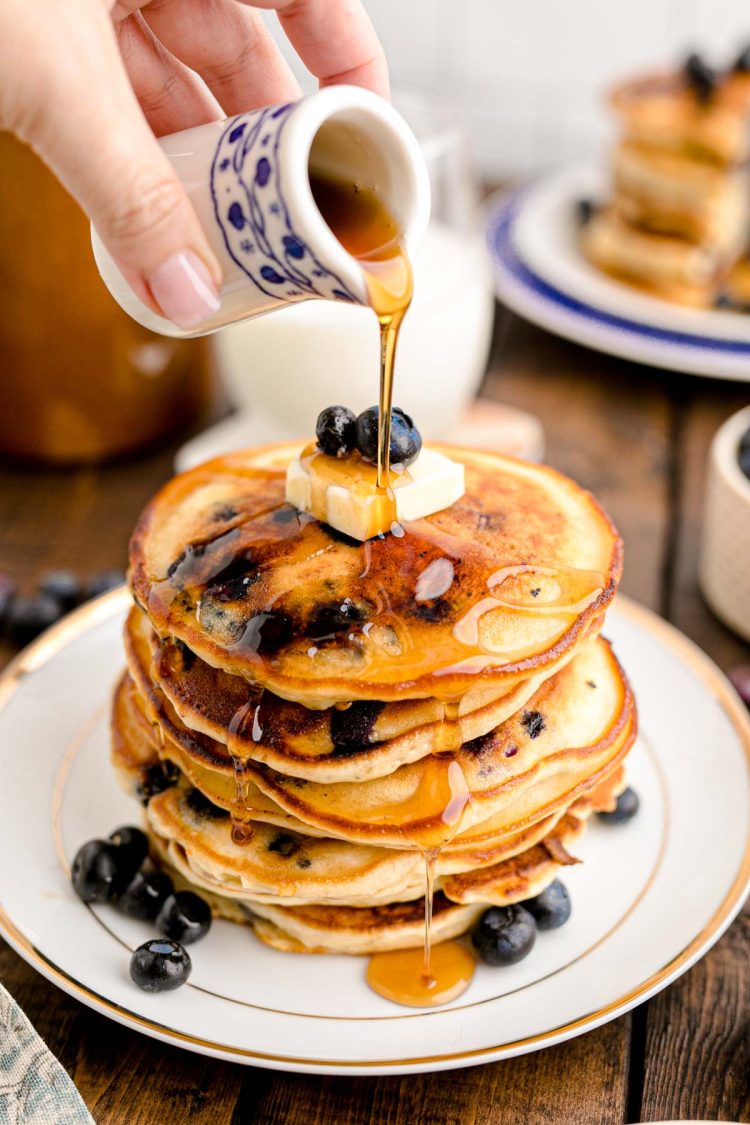 Close up photo of a woman's hand pouring maple syrup over a stack of blueberry pancakes on a white plate on a wooden table.