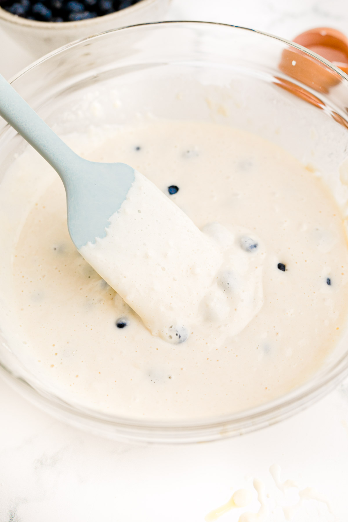Blueberry pancake batter in a glass mixing bowl with a blue rubber spatula.