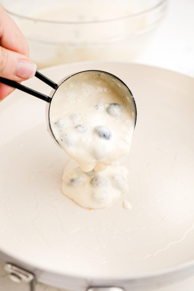A measuring cup pouring blueberry pancake batter onto a white skillet.
