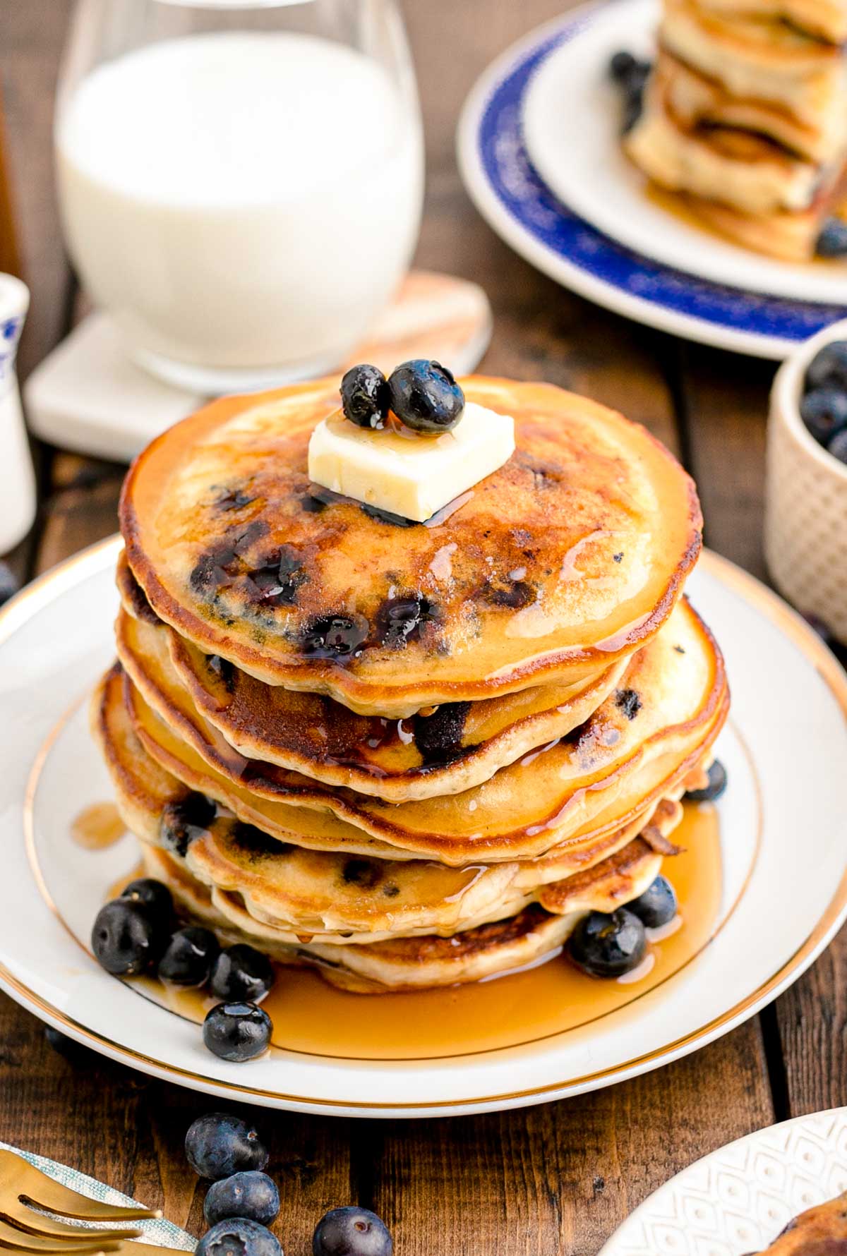 A stack of blueberry pancakes on a white plate on a wooden table with a glass of milk in the background. 