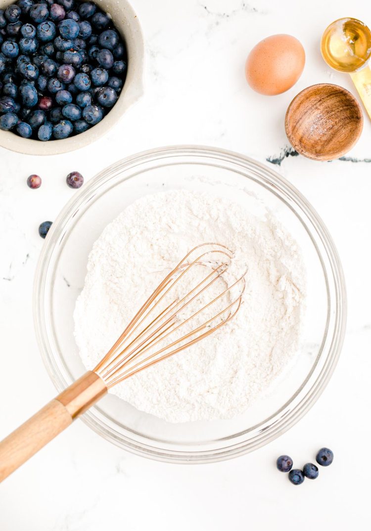 Overhead photo of dry ingredients being mixed in a glass bowl to make blueberry pancakes on a white table.