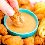 Close up photo of a woman's hand dipping a chick fil a nugget into homemade chick fil a sauce in a teal bowl.