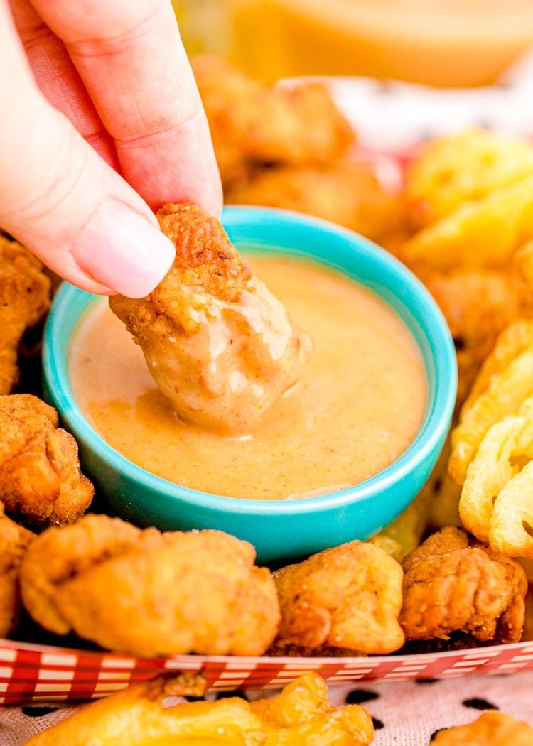 Close up photo of a woman's hand dipping a chick fil a nugget into homemade chick fil a sauce in a teal bowl.