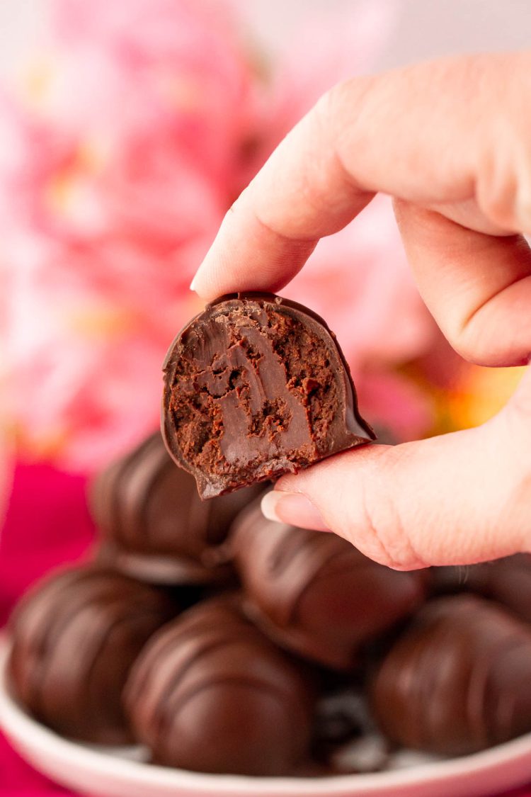 Close up photo of a woman's hand holding a chocolate truffle with a bite taken out of it. More truffles on a plate in the background.
