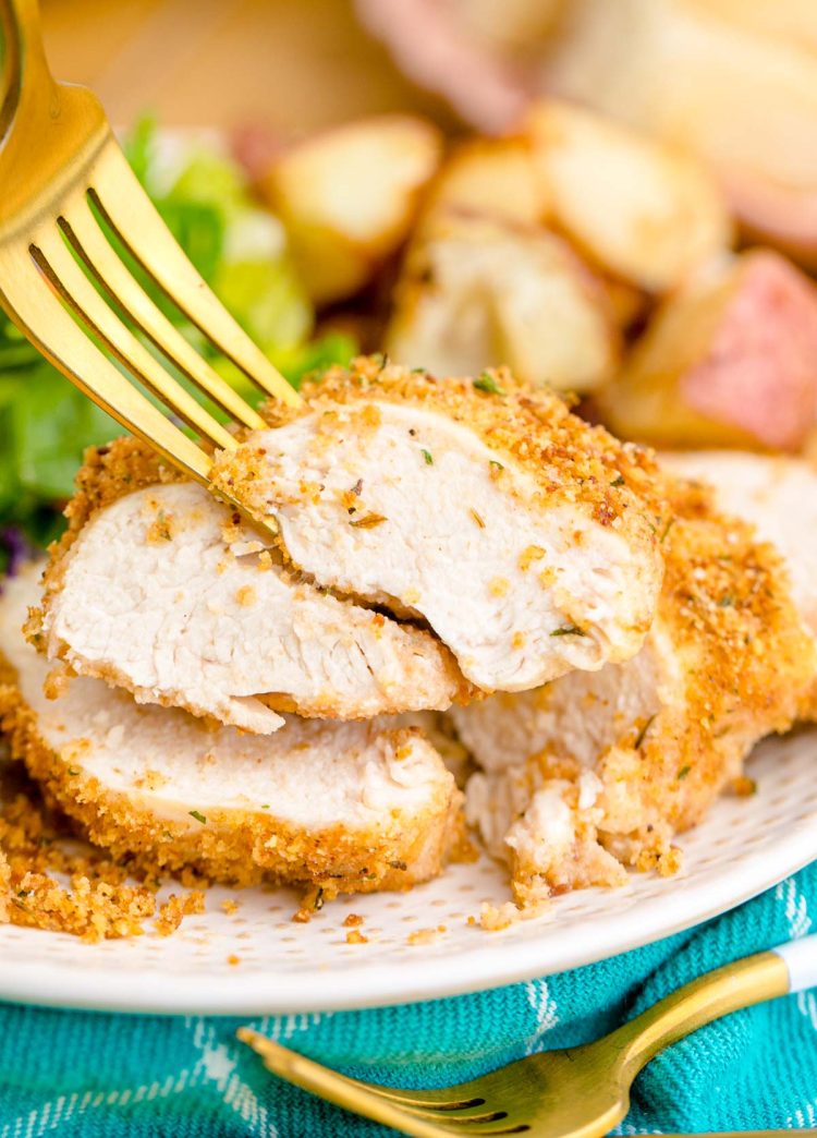 A fork taking a piece of shake and bake chicken off of a white plate.