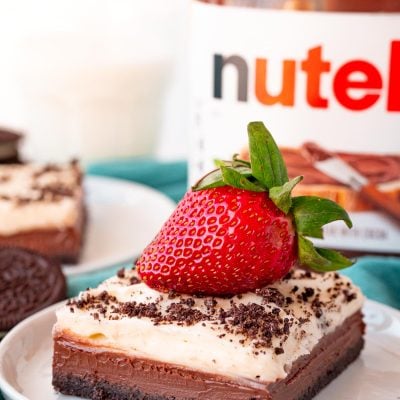 Close up photo of a slice of Nutella Cheesecake Bars on a white plate topped with a strawberry on a blue napkin.