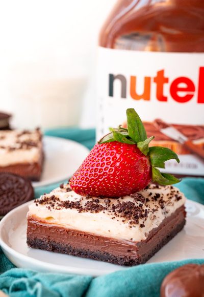 Close up photo of a slice of Nutella Cheesecake Bars on a white plate topped with a strawberry on a blue napkin.