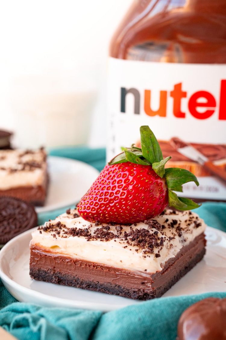 Close up photo of a slice of Nutella Cheesecake Bars on a white plate topped with a strawberry on a blue napkin.