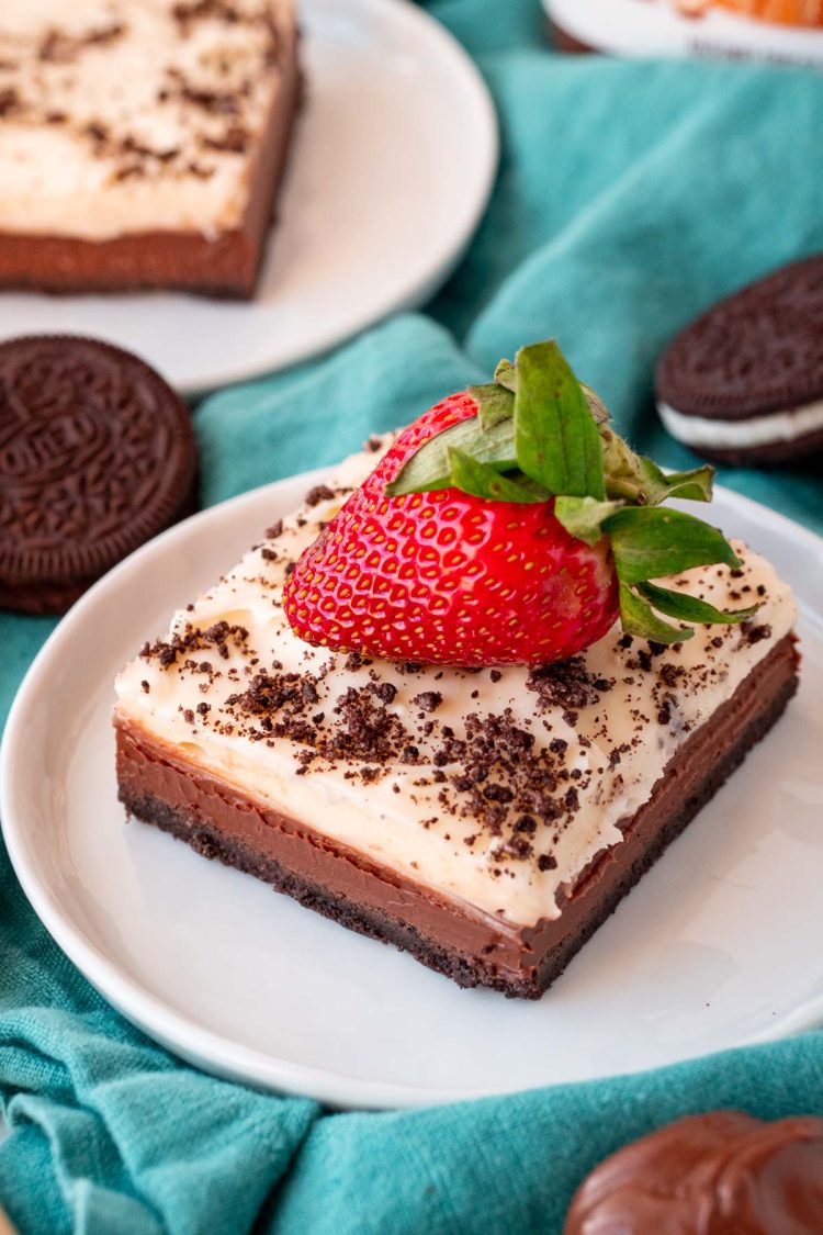 Close up photo of a slice of Nutella Cheesecake Bars on a white plate topped with a strawberry on a blue napkin.