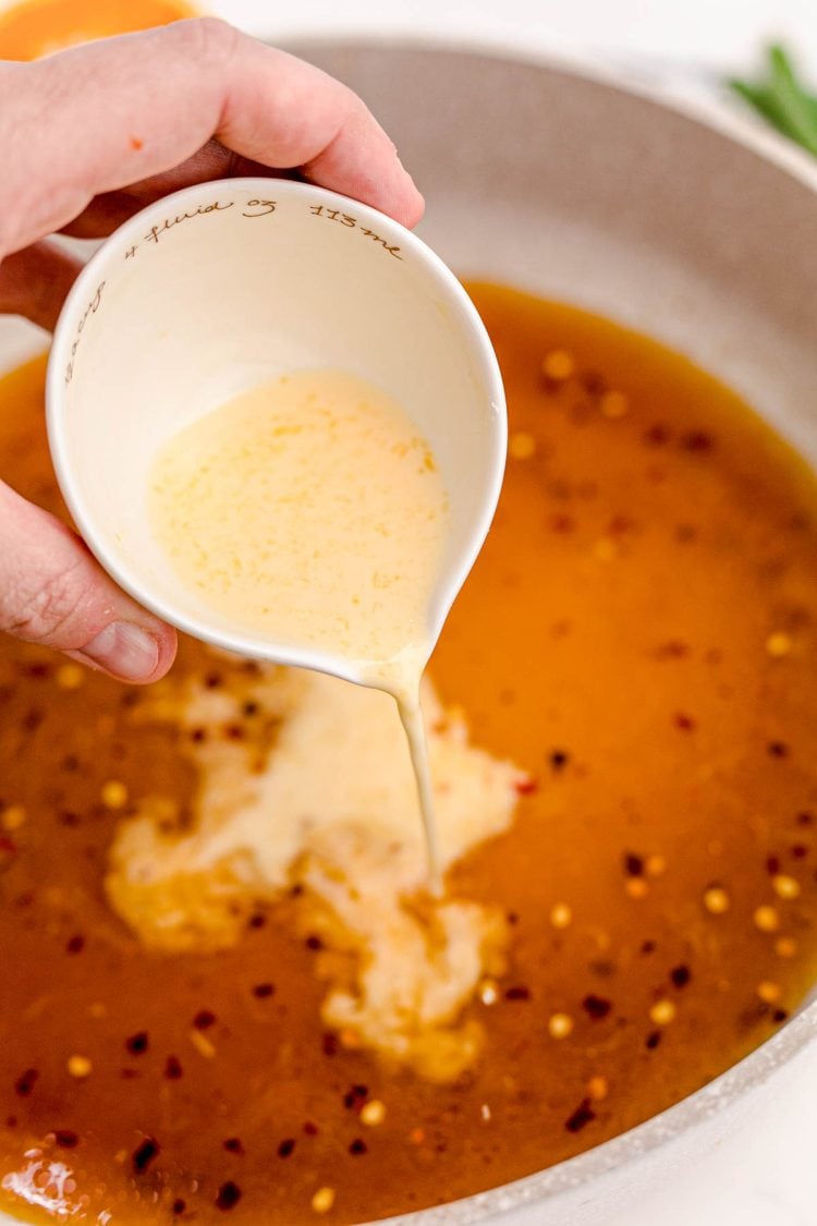 Cornstarch slurry being poured into a skillet with ingredients to make orange sauce.