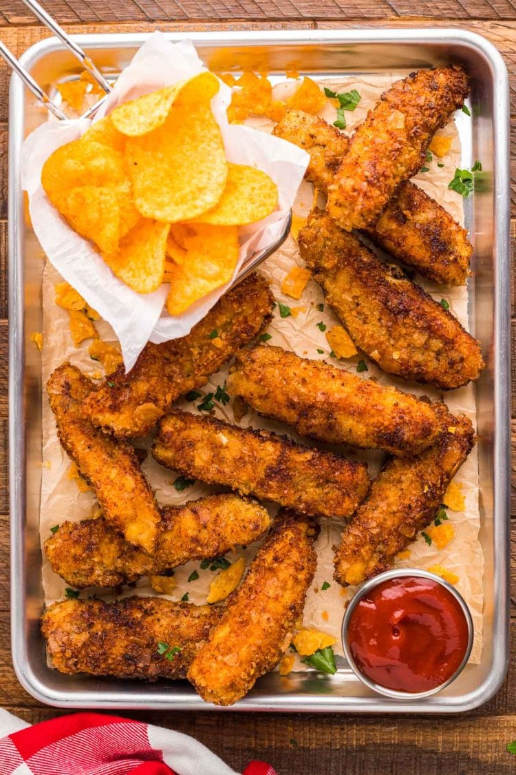 Overhead photo of potatoe chip chicken tenders on a metal serving tray with chips and sauce.