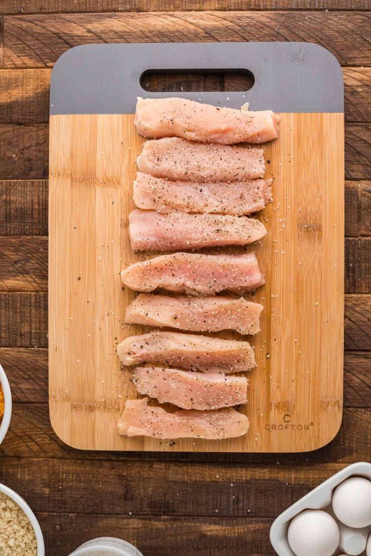 Strips of chicken breast being seasoned with salt and pepper on a cutting board.