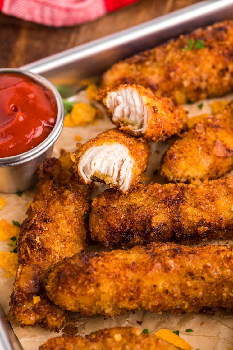 Potato chip breaded chicken tenders on a tray with one chicken tender broken in half to see the texture of the inside.