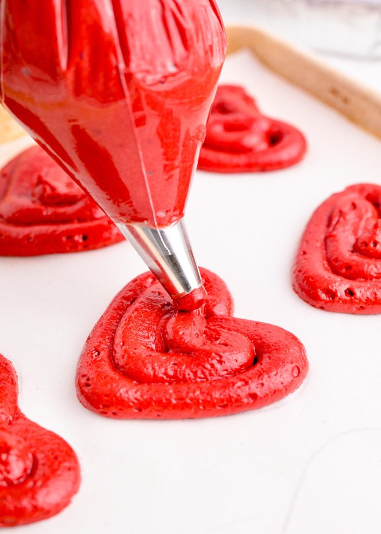 Red velvet cake batter being piped into heart shapes on a baking pan to make whoopie pies.