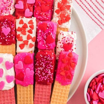 Close up photo of chocolate dipped wafer cookies decorated for Valentine's Day on a white plate on a pink surface.