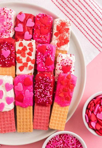 Close up photo of chocolate dipped wafer cookies decorated for Valentine's Day on a white plate on a pink surface.