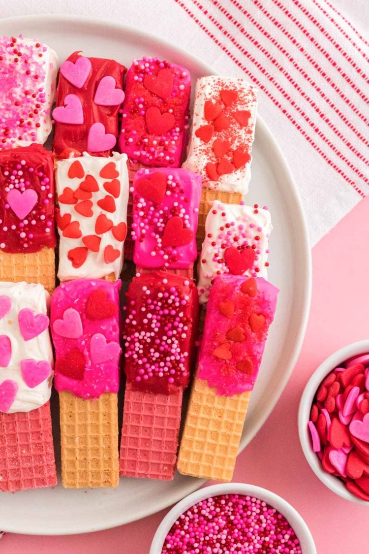 Close up photo of chocolate dipped wafer cookies decorated for Valentine's Day on a white plate on a pink surface.
