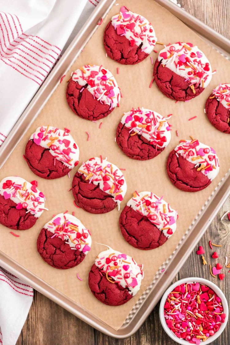 White chocolate dipped red velvet cookies on a parchment lined baking sheet with sprinkles for valentine's day.