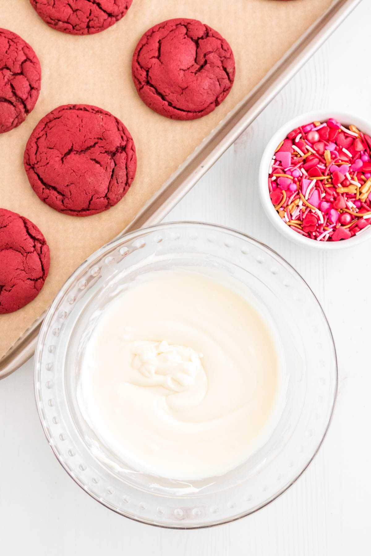 A bowl of melted white chocolate next to a baking sheet with red velvet cookies on it and a small bowl of sprinkles.